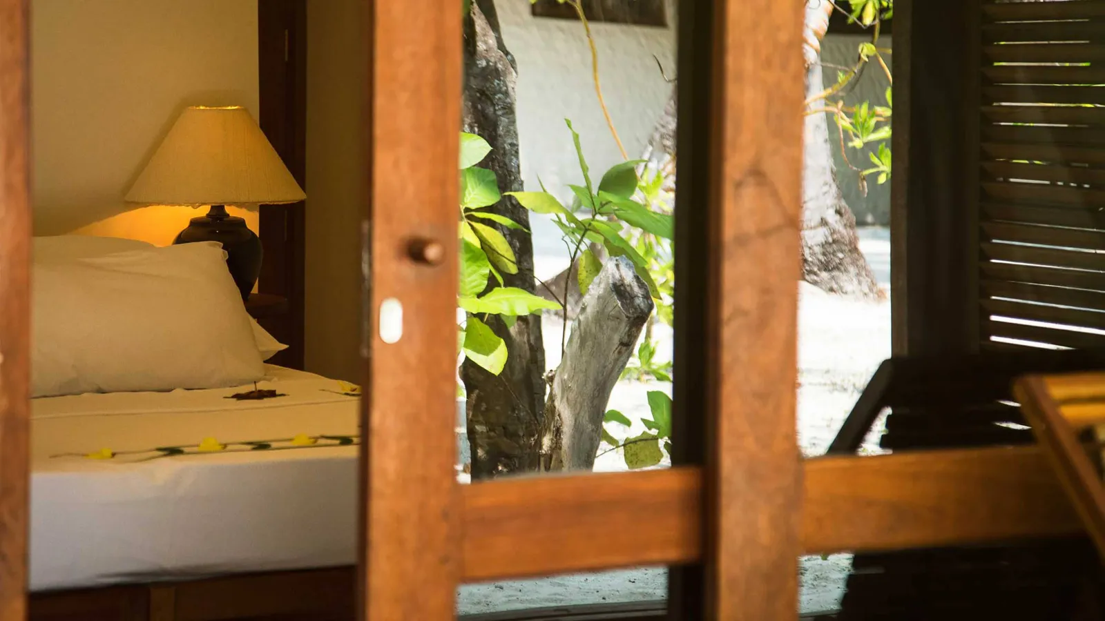 A garden villa seen through the French window: the bed, a lamp lit and the garden’s green beyond the shutters.