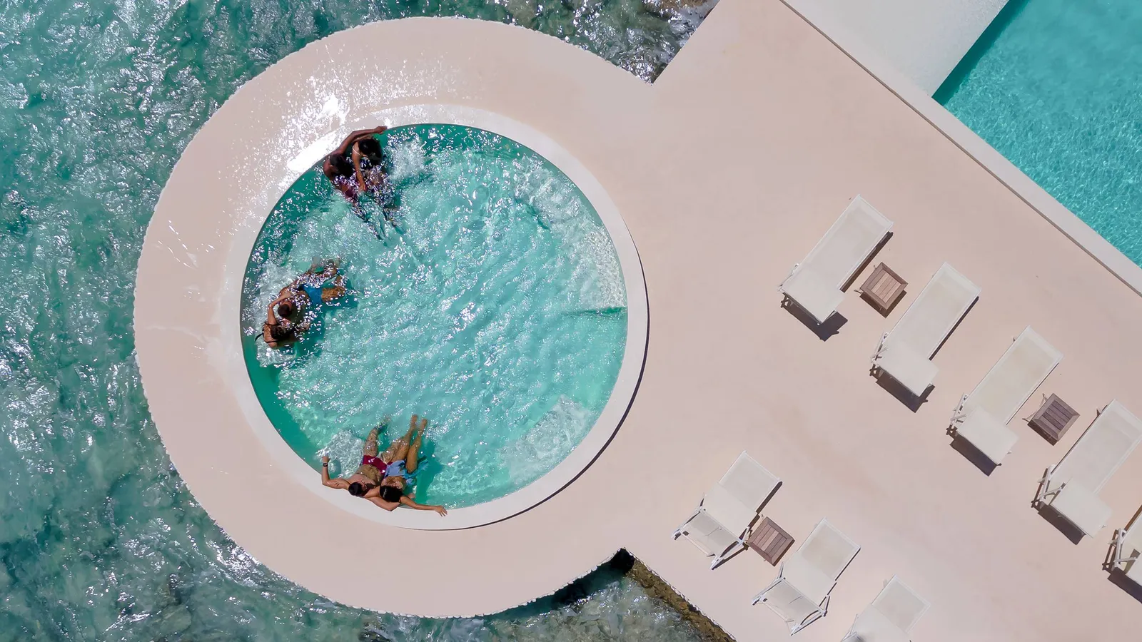 From above, the round whirlpool built over the sea, 3 guests in the water, beside the loungers of the infinity pool.