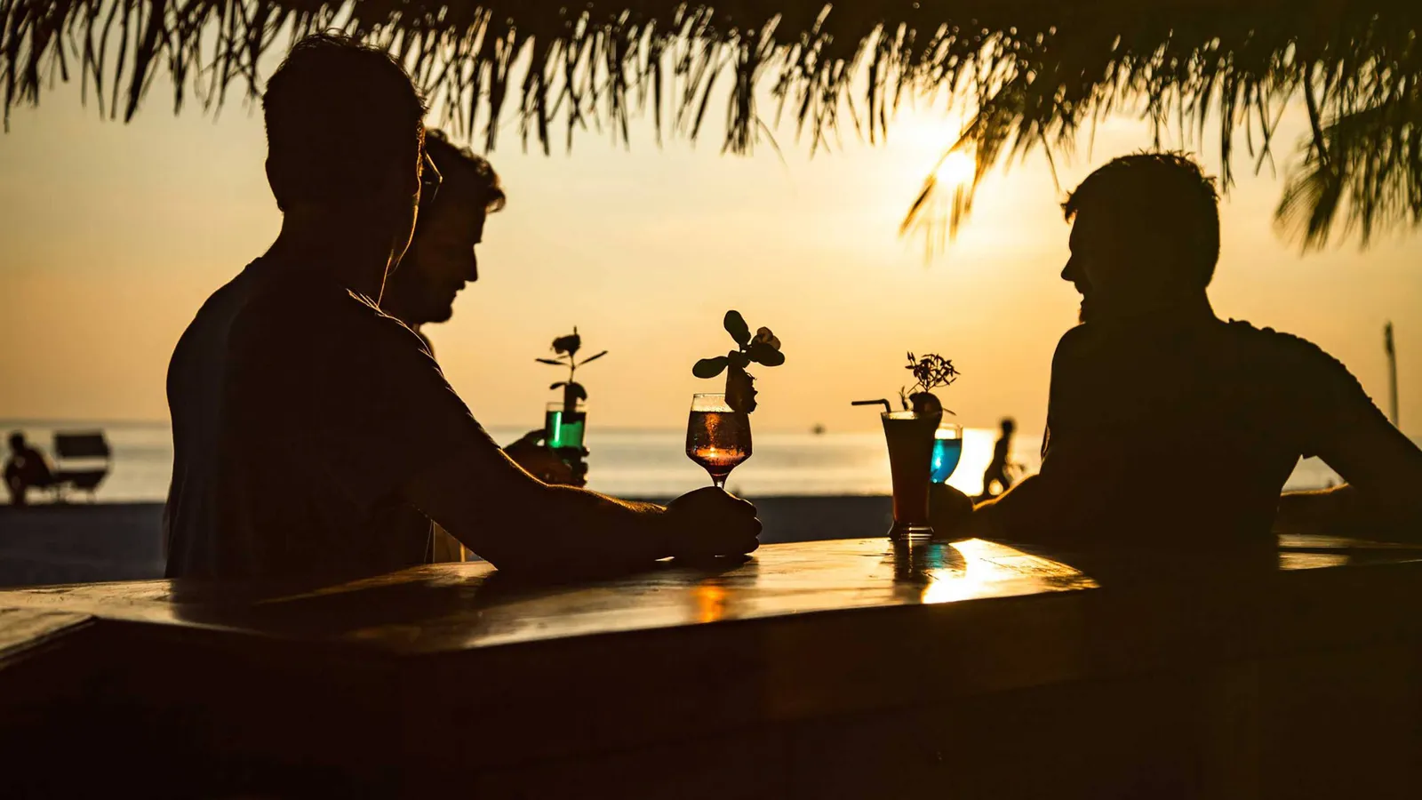 3 guests in silhouette at the Sunset Bar counter, with cocktails and the sun setting over the sea.