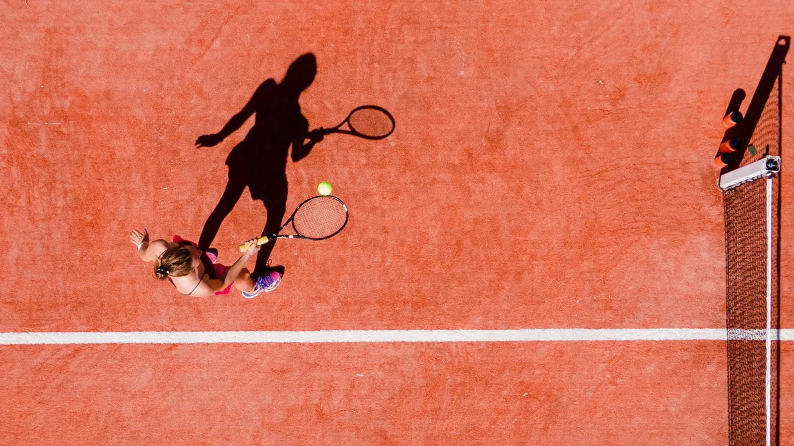 The red-clay tennis court seen from above, 2 players and their long shadows.