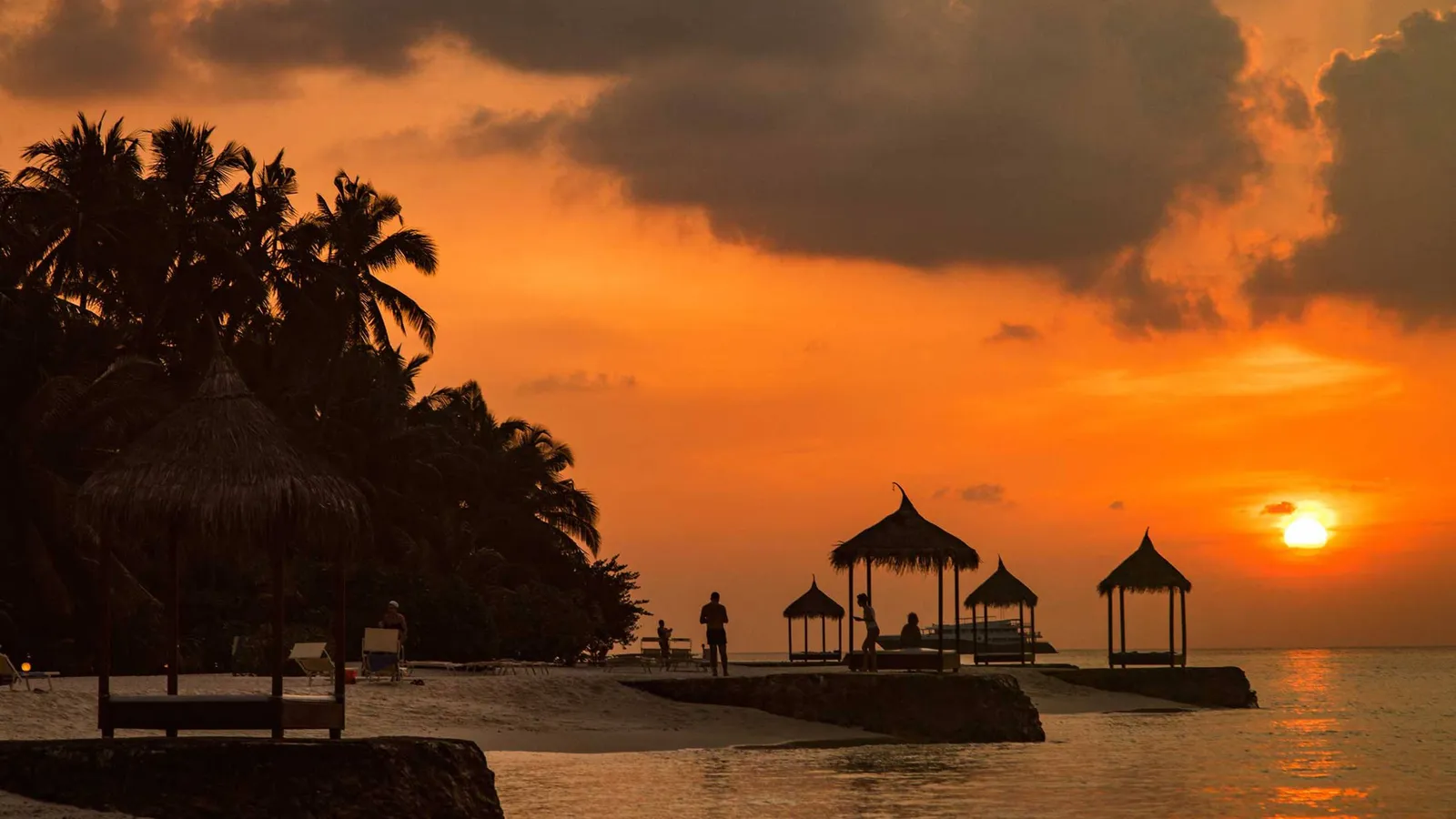 An orange sunset behind the thatched gazebos on the jetty, the palms in silhouette and a few guests on the shore.