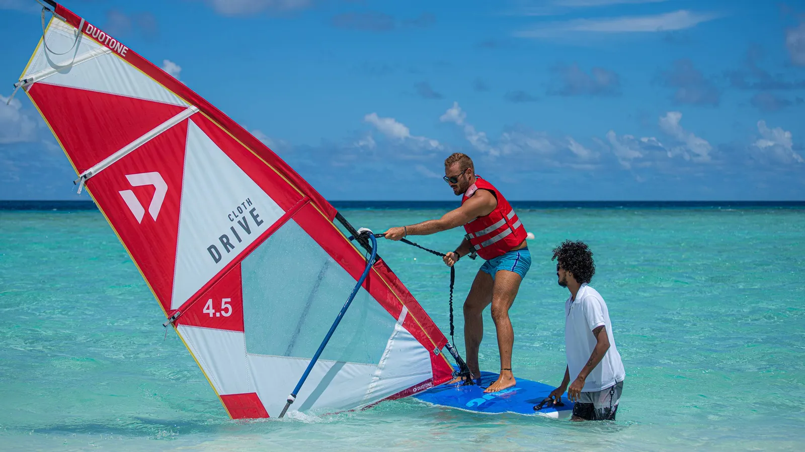 A guest tries windsurfing in the shallow lagoon, an instructor beside him.