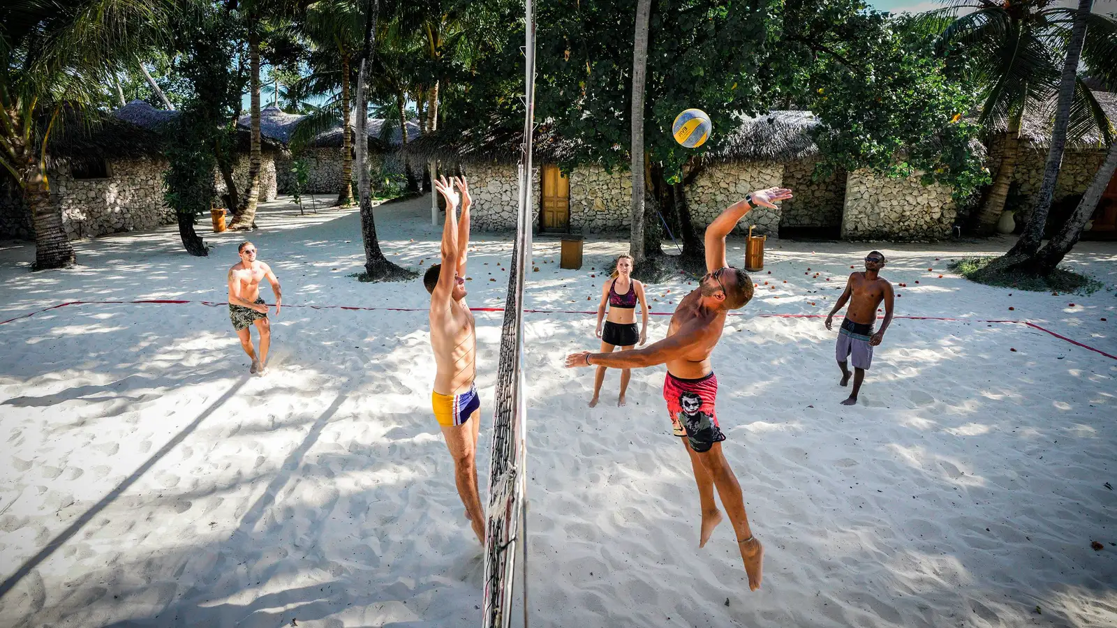 A beach volleyball game on the sand, among the palms and the coral-clad bungalows.