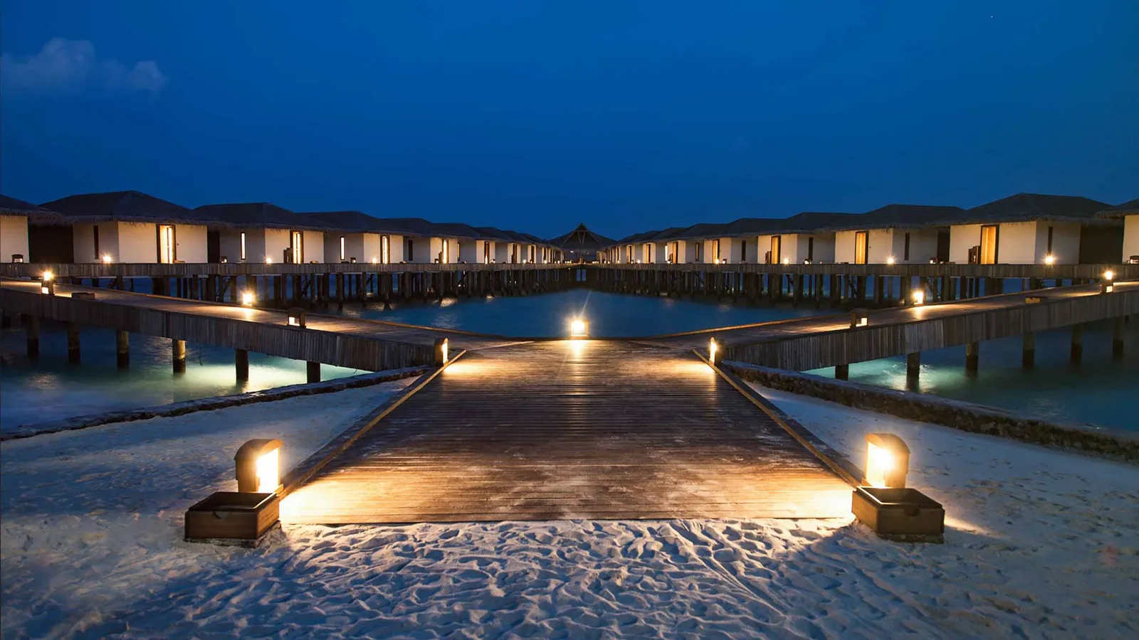 The jetty lit at dusk, with the water bungalows’ lights and the sand in the foreground.