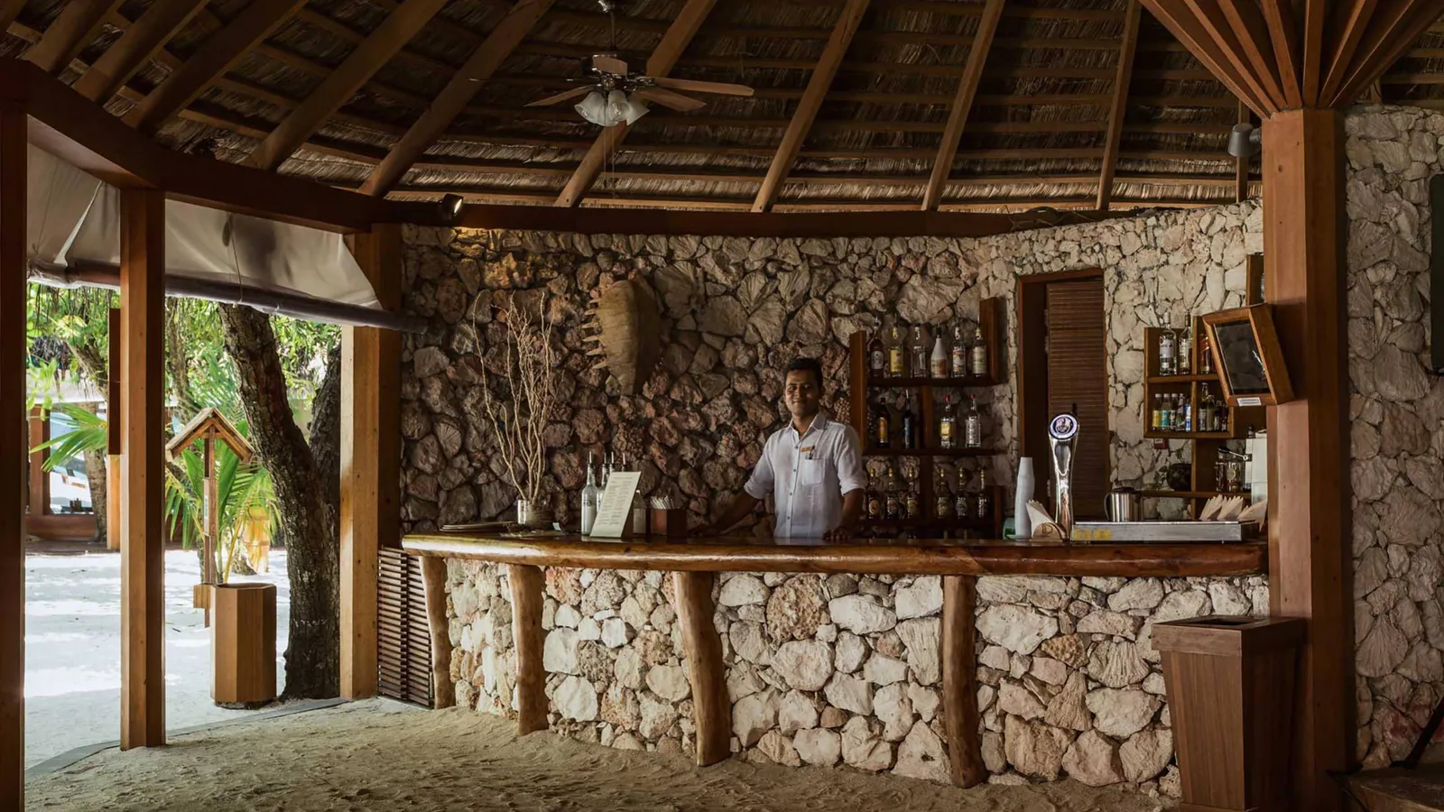 The main bar’s coral-clad counter under the timber roof, with a barman.
