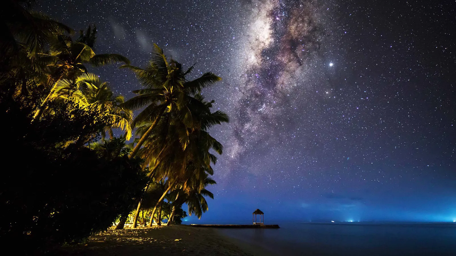 The Milky Way above lit palms, with a jetty and the dark sea on the horizon.