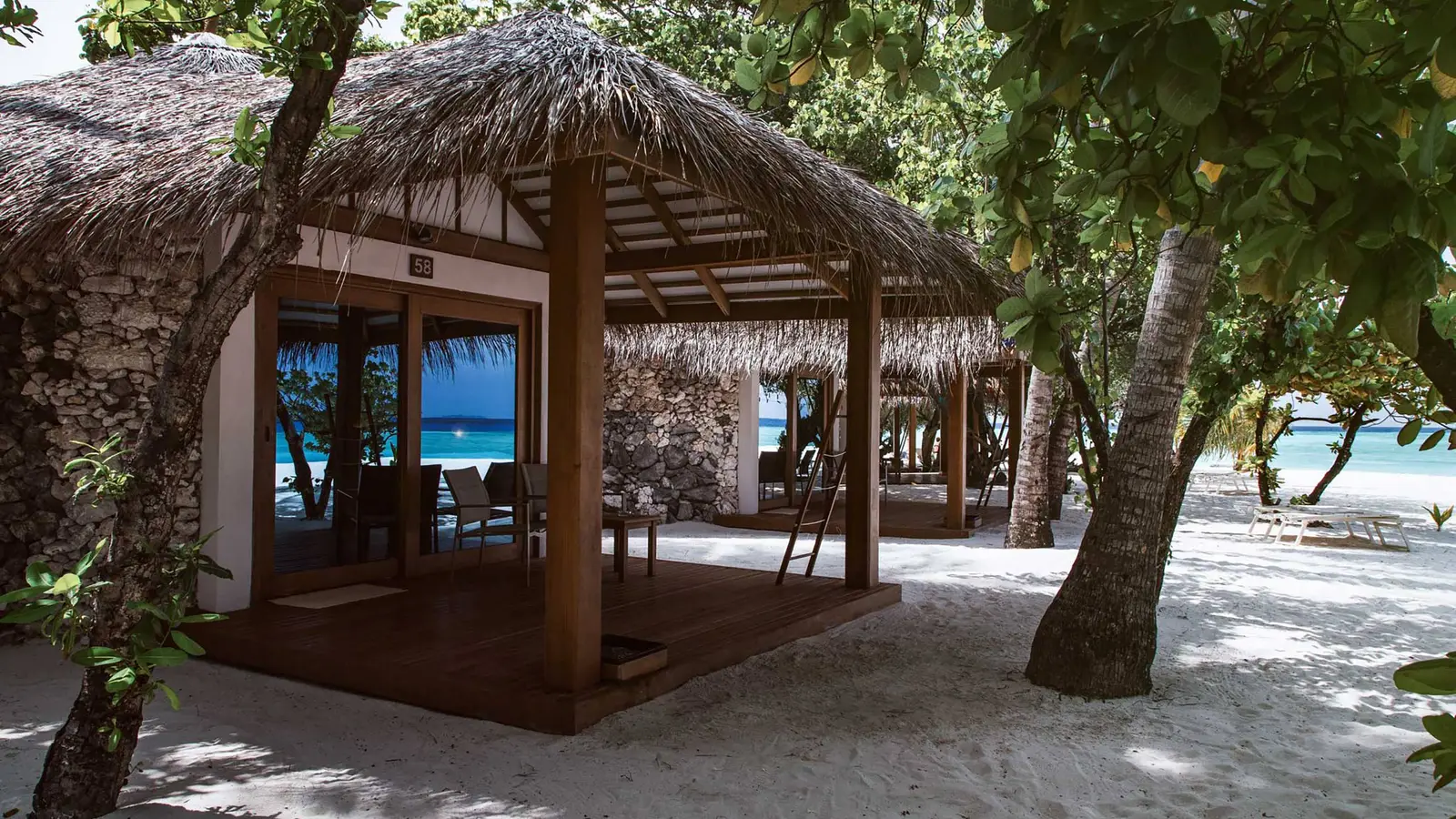 A beach bungalow with a thatched roof and a timber veranda, among the trees on the sand.