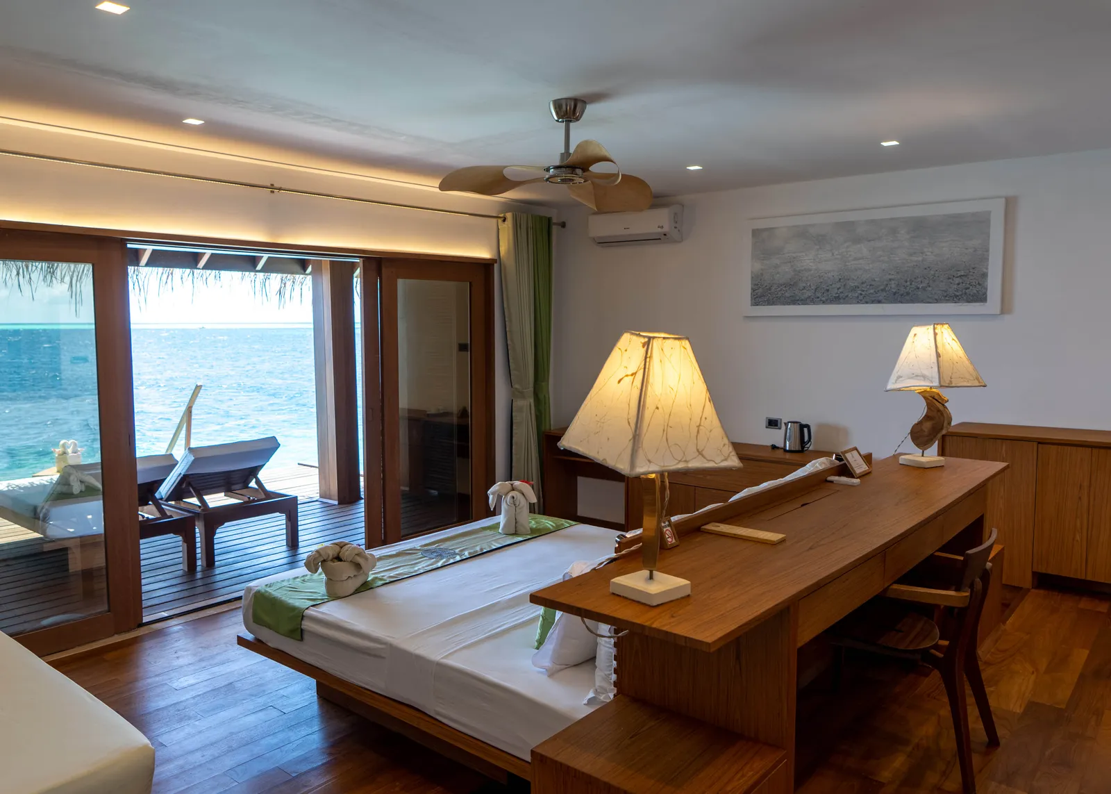 A water suite: the bed, the timber desk and the glass door open onto the deck and the sea.
