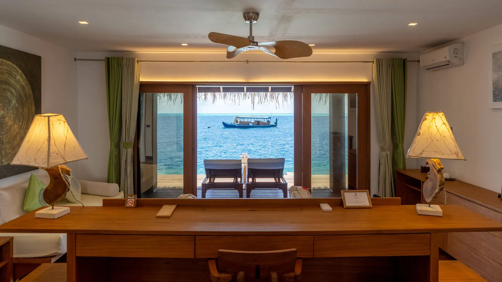 The water suite’s desk facing the glass doors, 2 loungers outside and a dhoni on the sea.