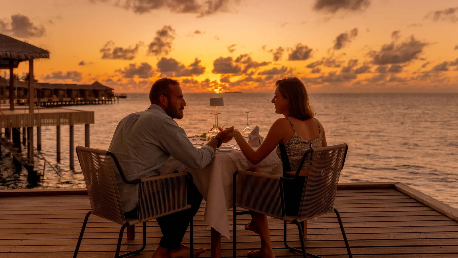 A couple dine on a timber deck at sunset, the water bungalows behind them.