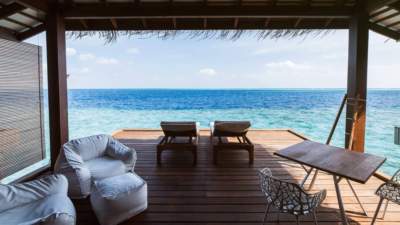 A water bungalow’s deck seen from inside: 2 loungers, the armchairs and the open ocean.