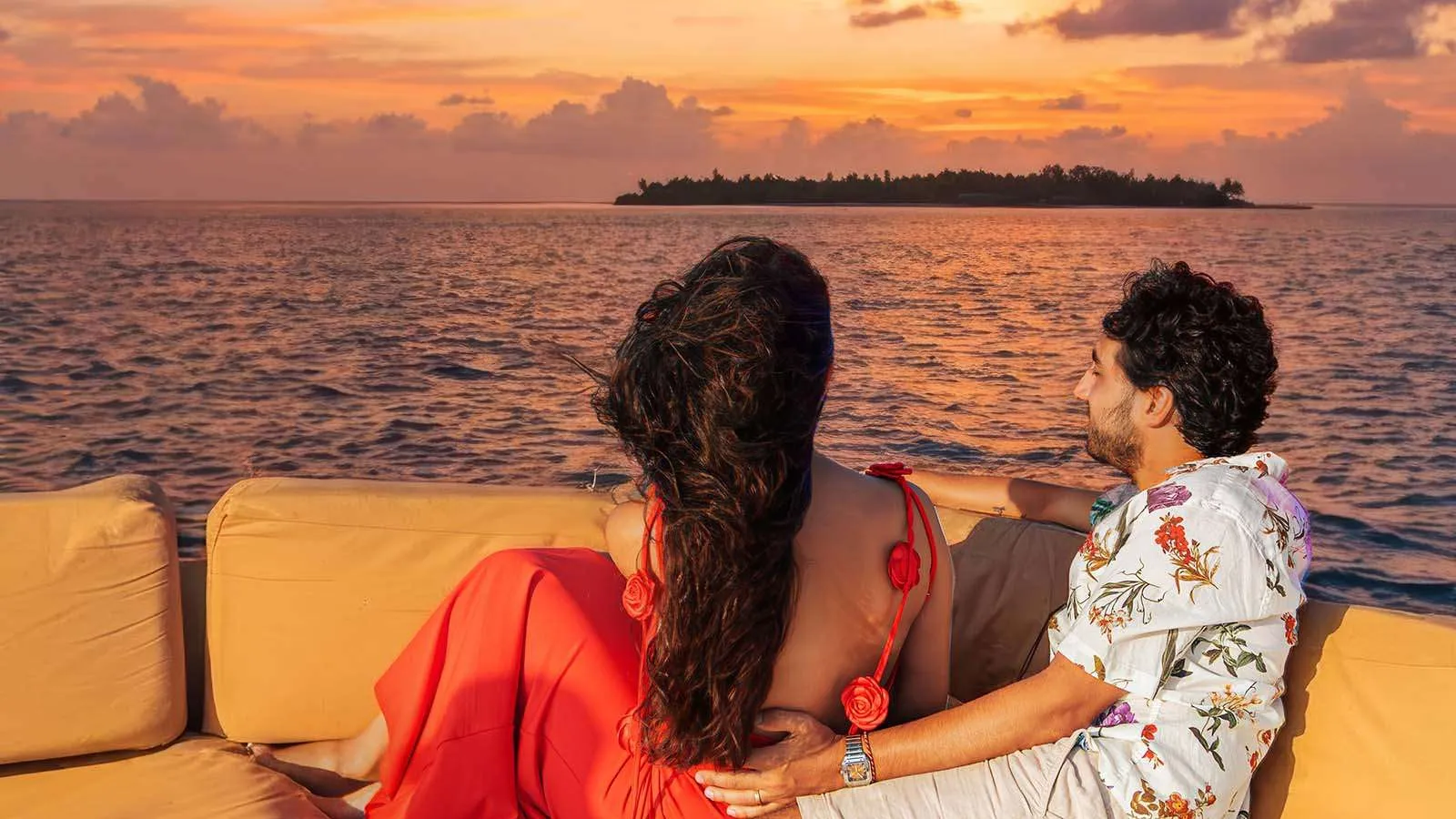 Una coppia sul divano di prua di una barca al tramonto, davanti al mare e a un’isola all’orizzonte.