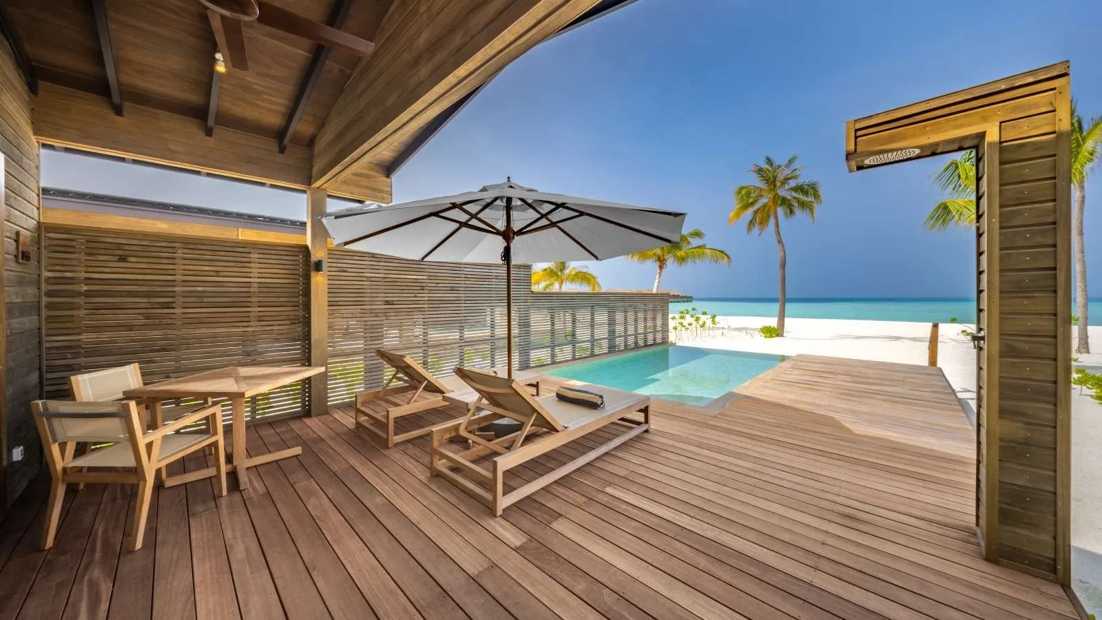 La terrazza coperta di una Sunset Beach Pool Villa: tavolo, lettini, ombrellone e la piscina verso la spiaggia.