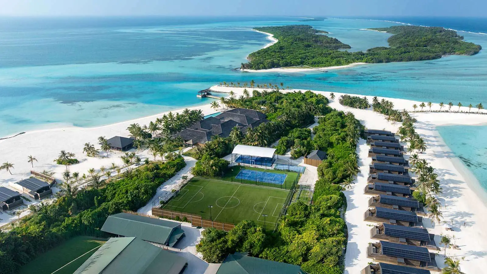 L’isola dall’alto: il campo da calcio, i campi da tennis e padel in mezzo al verde e le ville sulla spiaggia a destra.