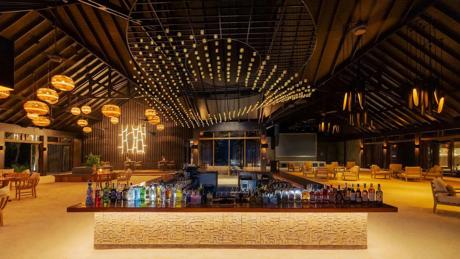 Il Velu Bar di sera: il lungo bancone illuminato, le bottiglie e i tavoli sotto il soffitto di luci.