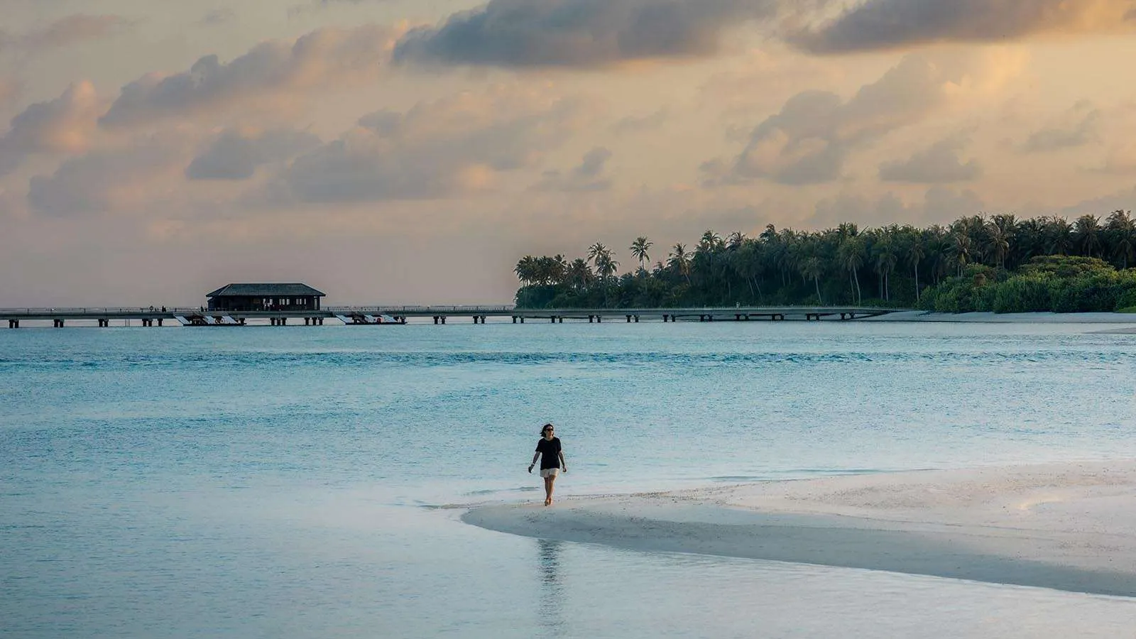 Una donna cammina sulla lingua di sabbia al crepuscolo, con la laguna calma e un pontile all’orizzonte.