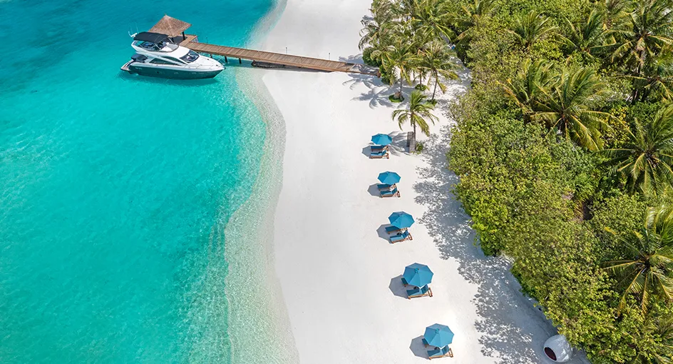 The arrival jetty from above, a boat moored alongside, the white beach and the row of blue parasols under the palms.