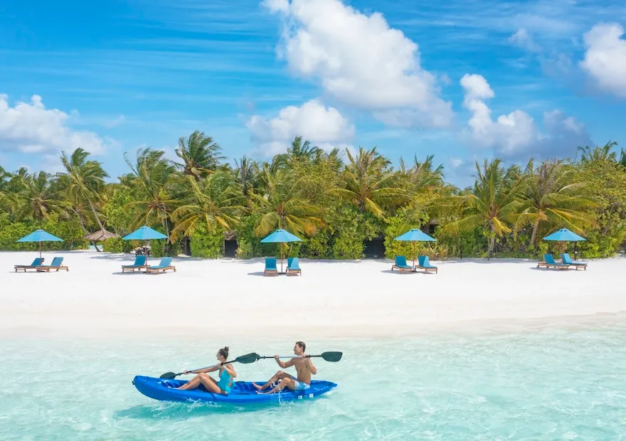 2 guests kayaking in the shallow lagoon off the beach, with its parasols and palms.
