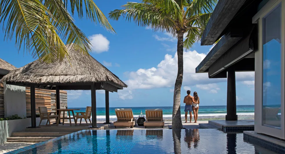 A couple at the edge of an Ocean House’s infinity pool looks out to the ocean, between the thatched pavilion and the palms.