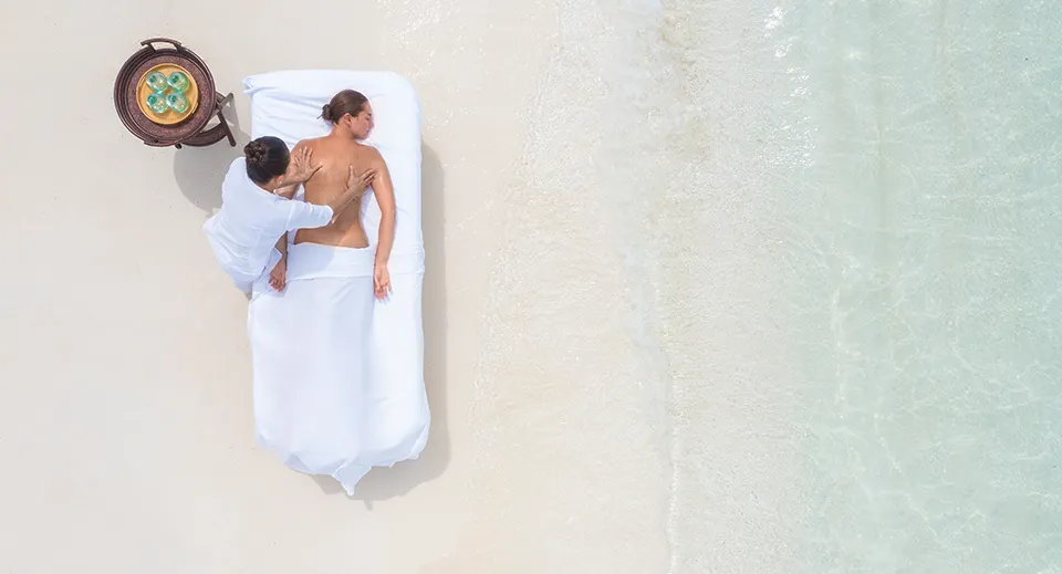 A massage on the white sand seen from above: a guest on the couch, the therapist beside her and the pale lagoon.