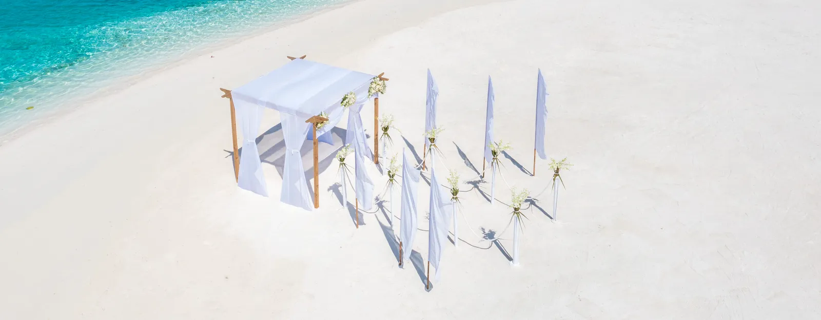 A ceremony laid out on the beach, seen from above: the white canvas gazebo, the flowers and the sand running into the lagoon.