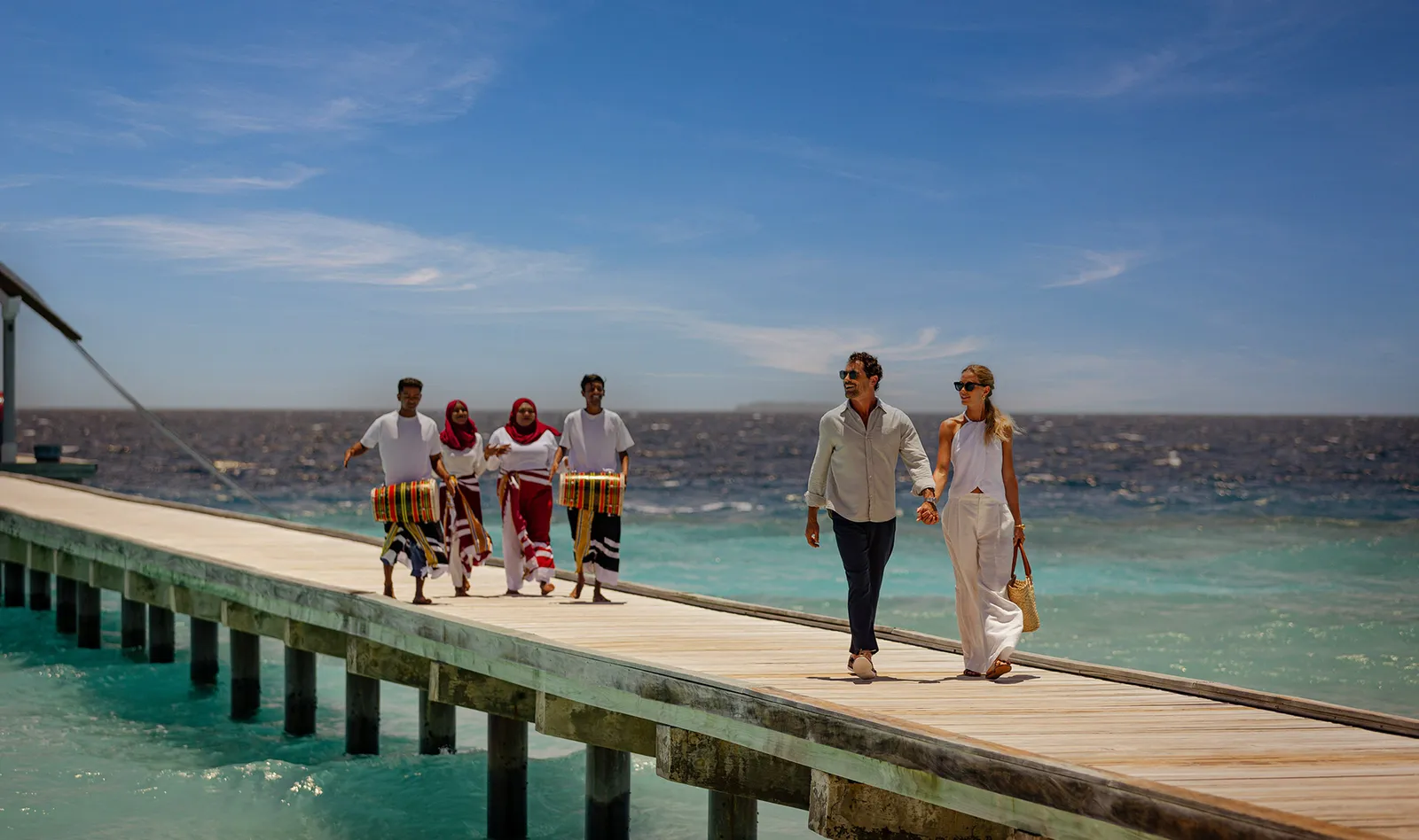 A couple walk along the arrival jetty, welcomed by 4 of the resort’s staff with drums.