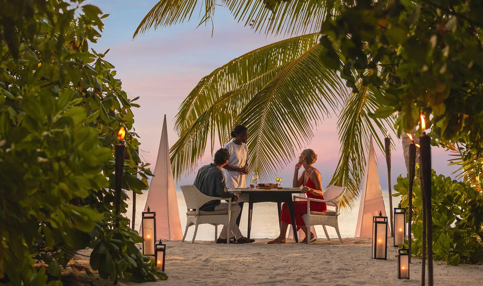 Dinner on the beach at dusk, among torches and lanterns, under a palm.