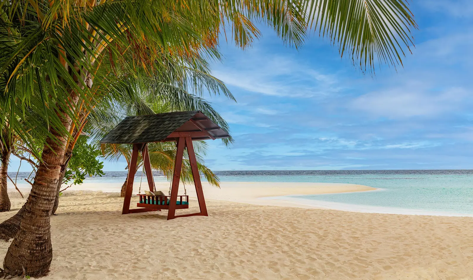A timber swing on the beach, under a thatched roof, facing the lagoon.