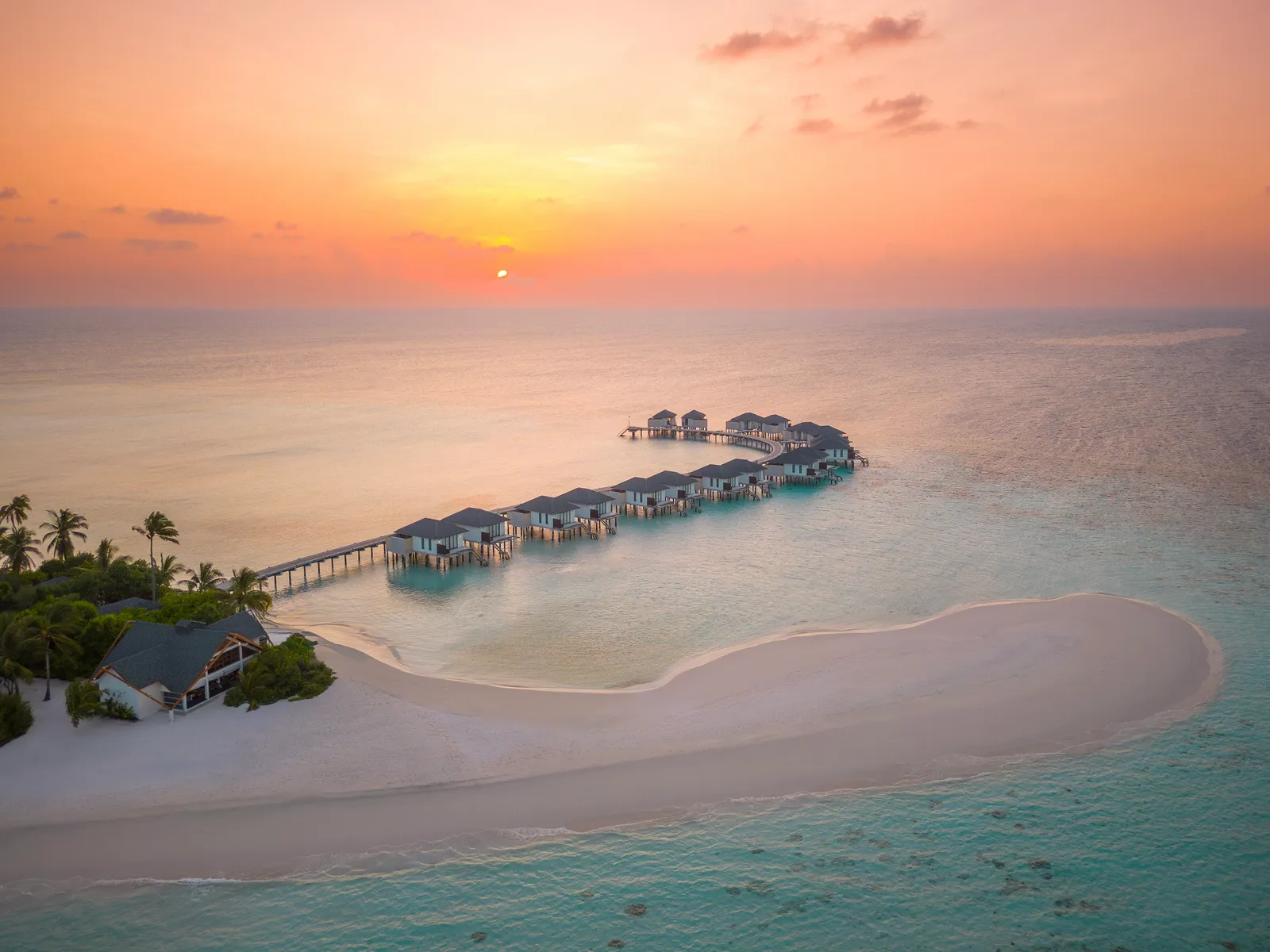 The water-villa jetty at sunset, seen from above, over the lagoon and the sandy tip of the island.