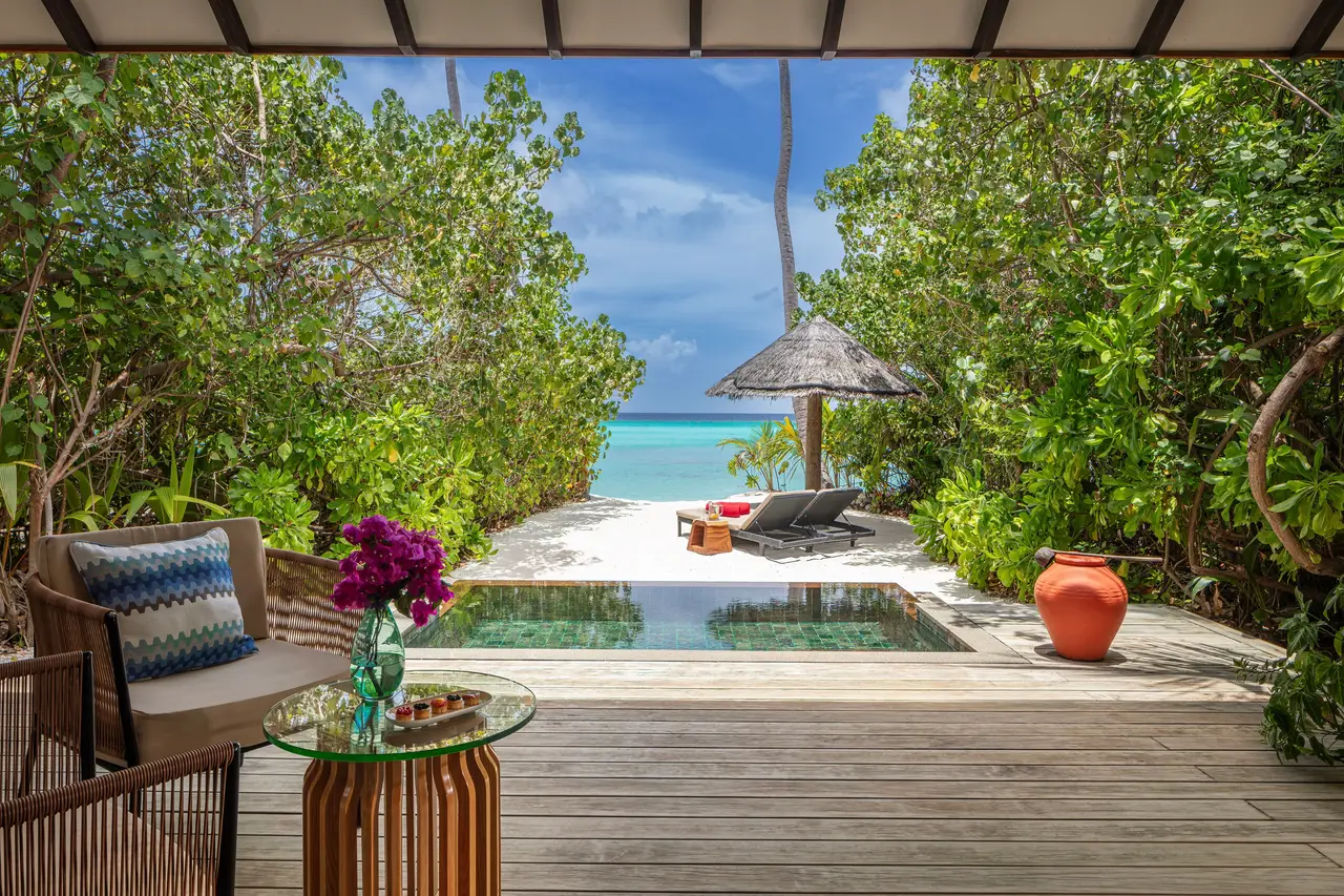 A sunset-side pool villa: the armchair, flowers on the table, the plunge pool and the beach beyond the green.