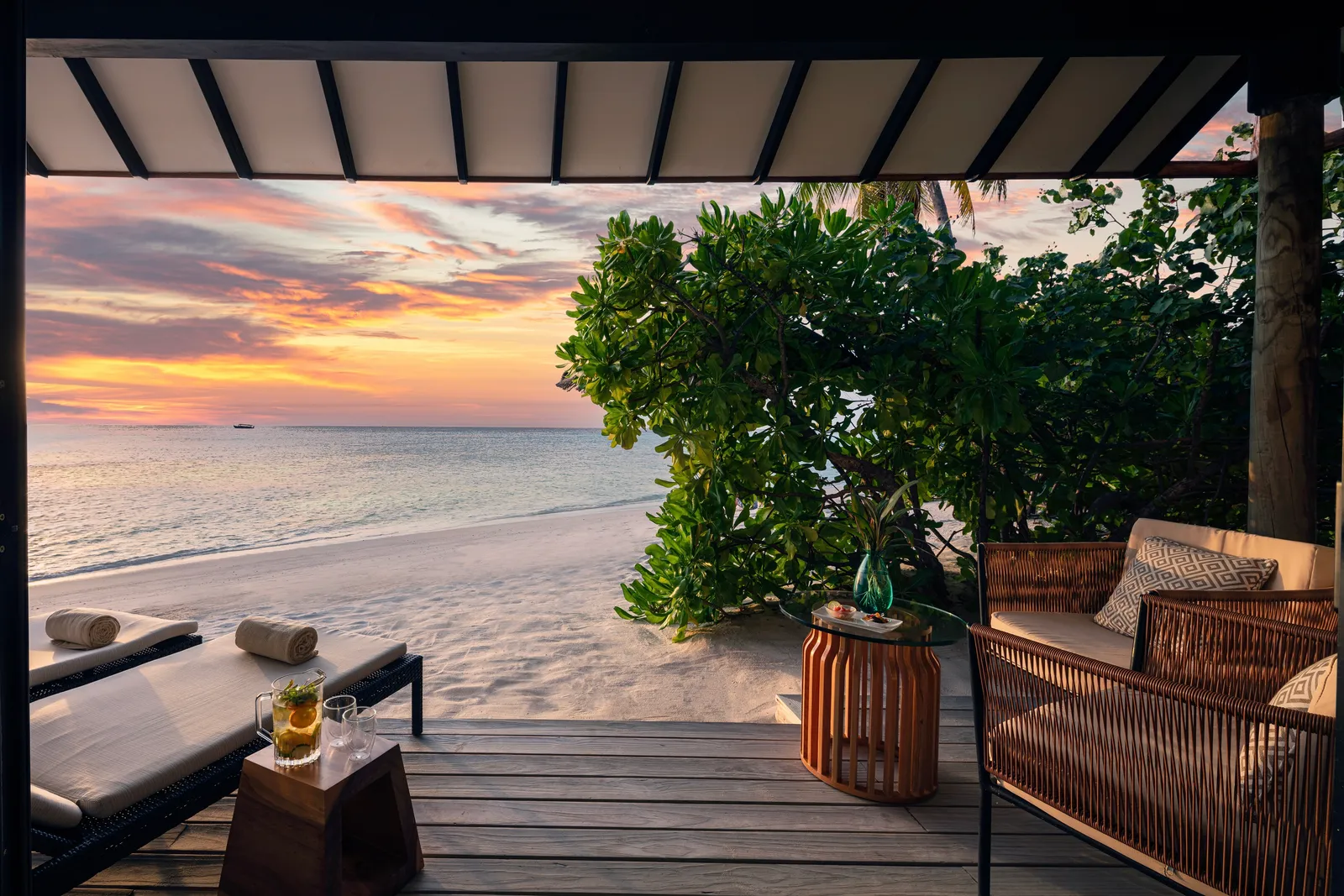 The deck of a sunset beach villa, with armchairs, loungers and an orange sky over the sea.