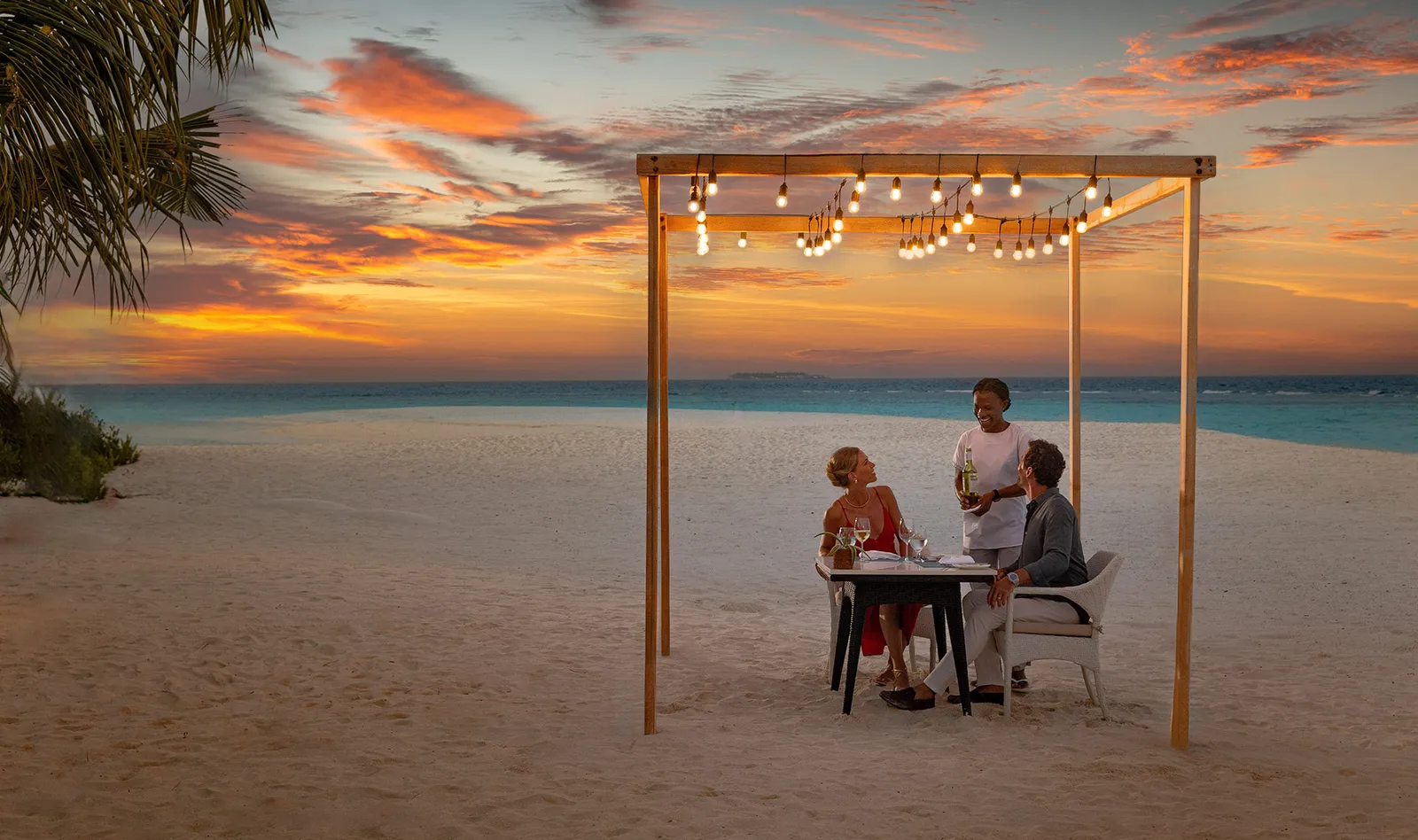 Dinner for 2 on a sandbank at sunset, under a frame of string lights, served by a waiter.