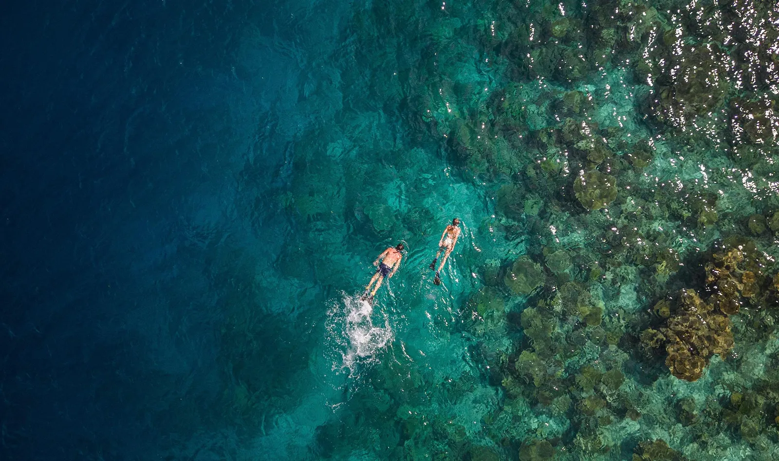 2 people snorkelling over the reef, seen from above, where the lagoon turns deep blue.