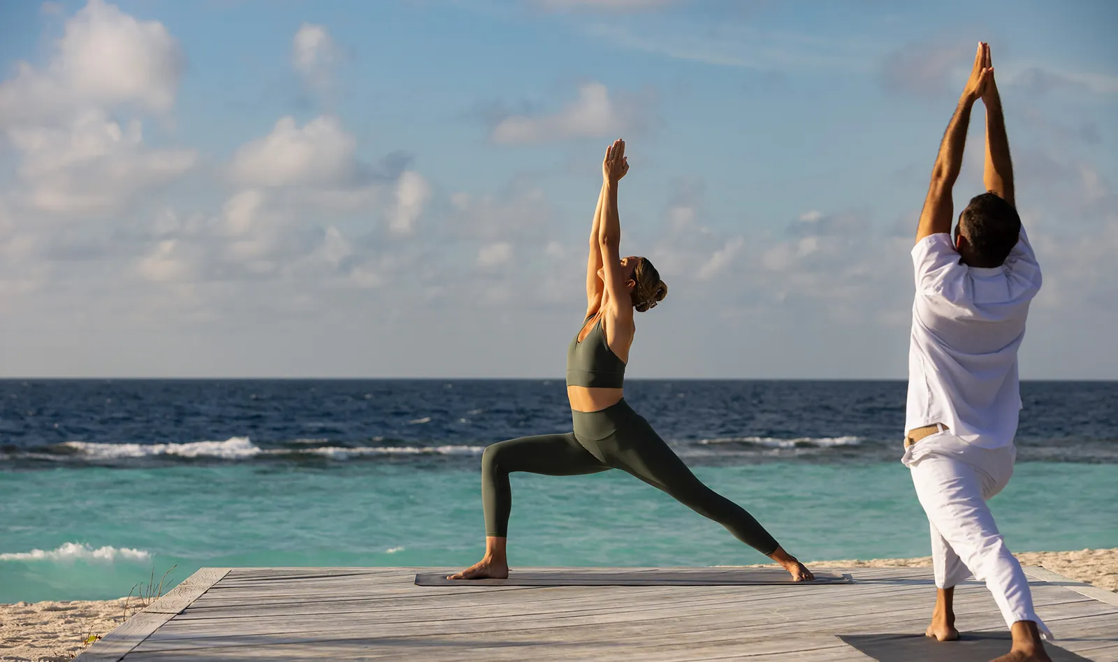 2 people doing yoga on a timber platform facing the sea.