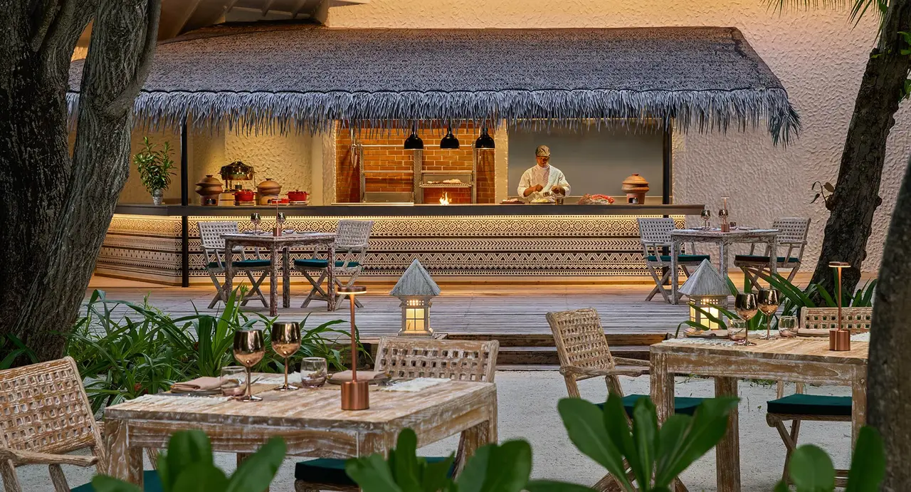Alifaan under its thatched roof: the chef at the open fire at the back, the timber tables and the lanterns on the sand.