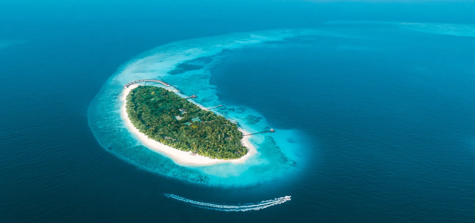 Fonimagoodhoo from above: the green island and its white beach inside a ring of reef and lagoon, with a speedboat leaving its wake.