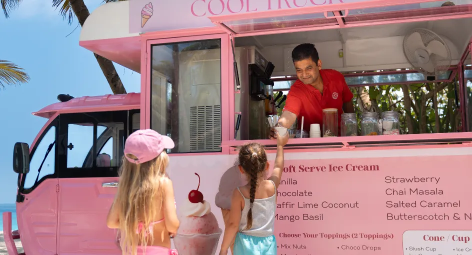 The pink ice-cream van under the palms, 2 girls at the counter and the server handing over a cone.