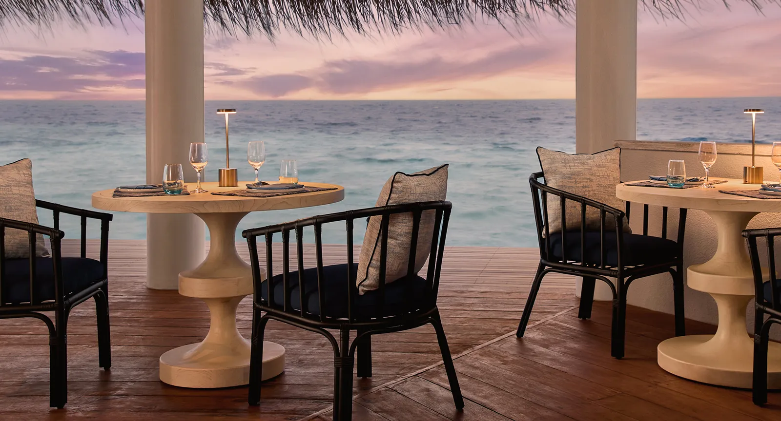 Kaiyo’s tables on the timber deck at dusk, the lamps lit, the dark chairs and the sea under a pink sky.
