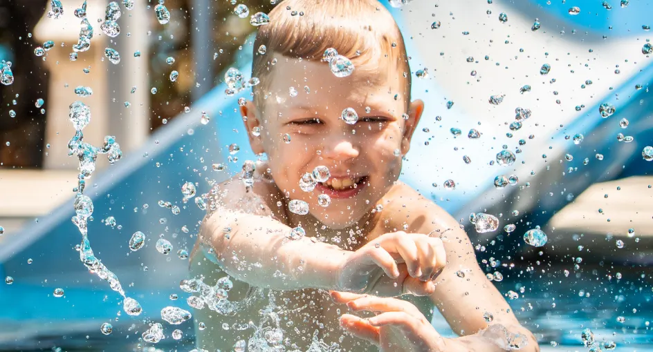A boy laughing in the spray at the bottom of a water slide, in the kids’ club pool.