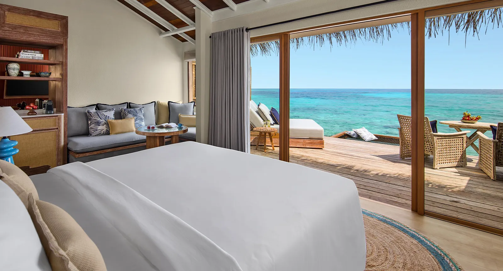 A water villa bedroom, the glass doors wide open onto the deck, the loungers and the turquoise lagoon.
