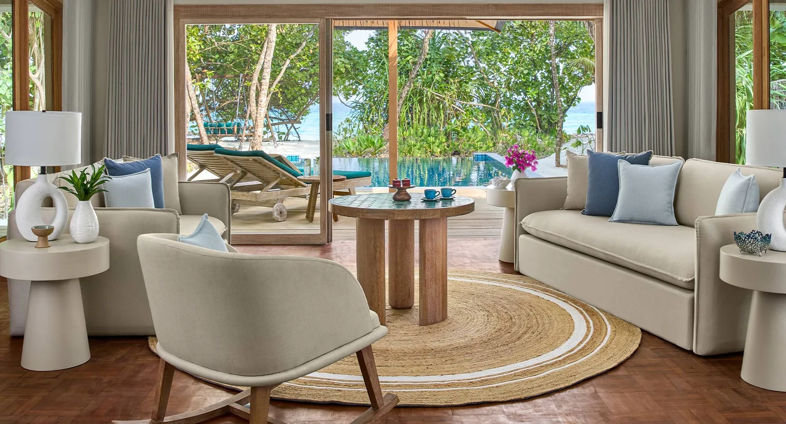 The Reethi Suite living room, its pale armchairs, rope rug and glass doors open onto the pool and the trees.