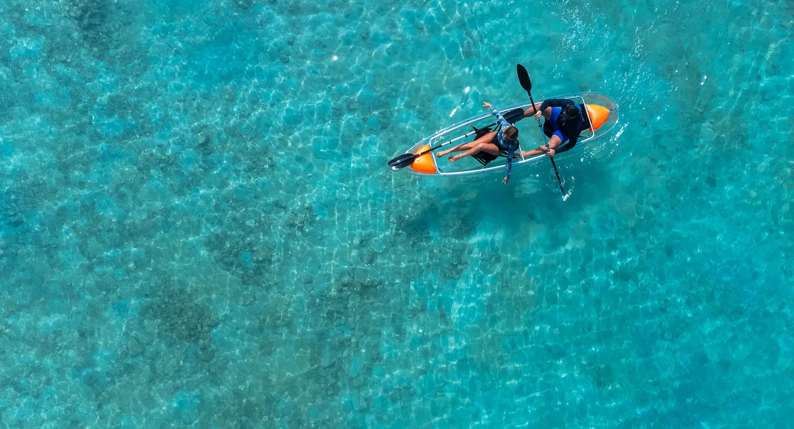 A couple in a clear canoe on the turquoise lagoon, seen from above.