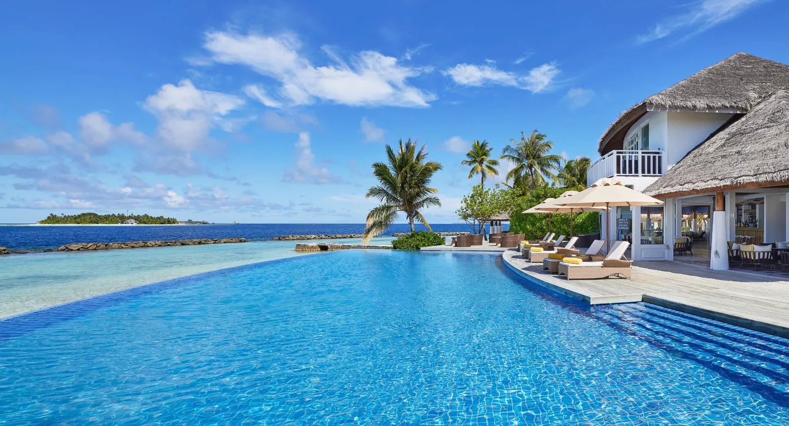 The infinity pool under a blue sky, its edge on the sea, with the palms and the loungers beside the bar.