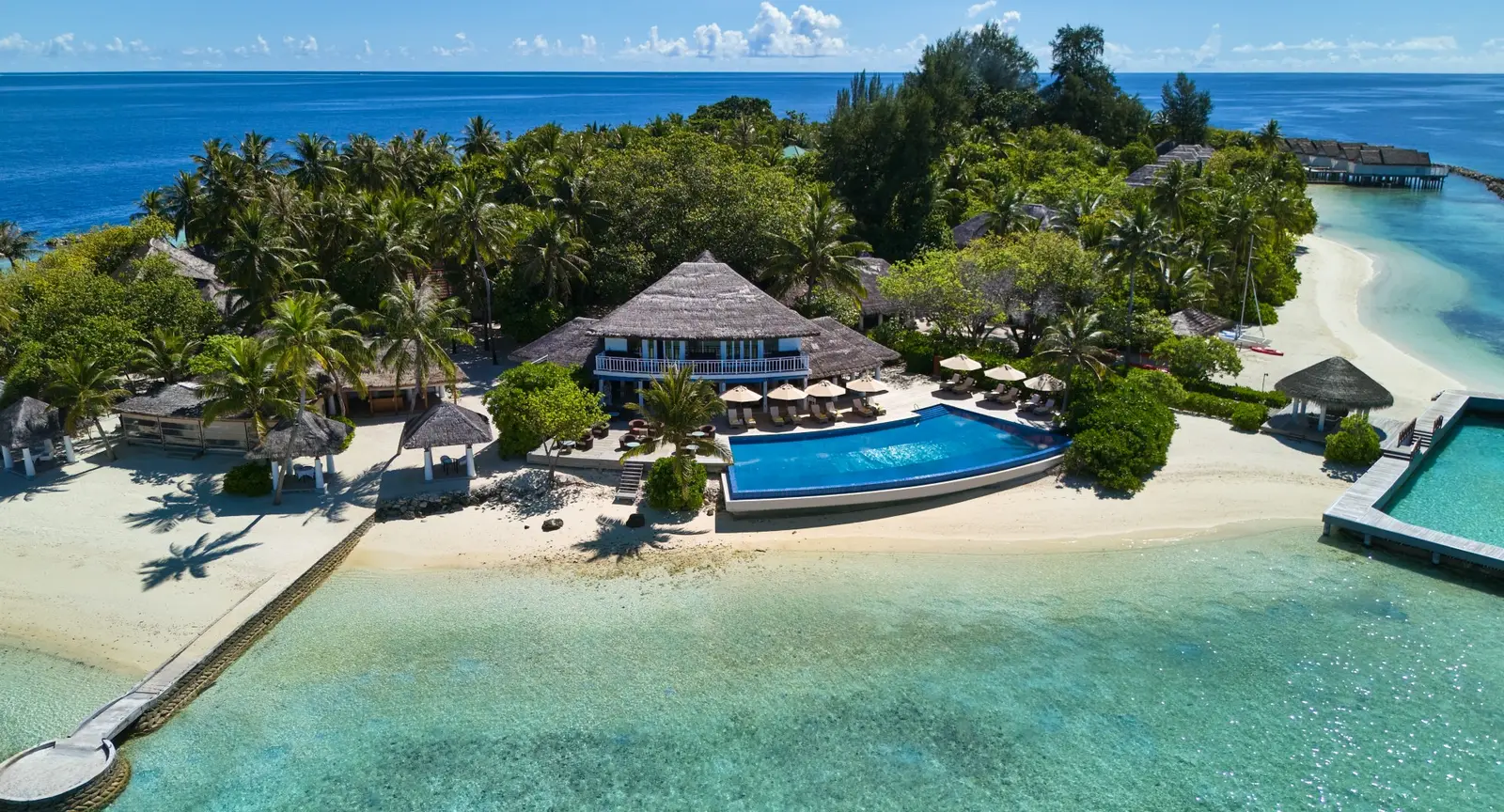 The main building under its thatched roof, the infinity pool on the beach, the parasols and the lagoon closed off by rocks.