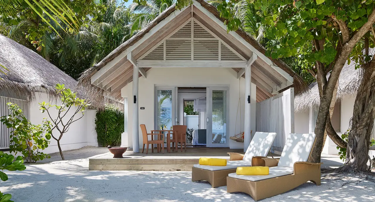 The front of a beach villa: the porch, loungers on the sand and the trees around it.
