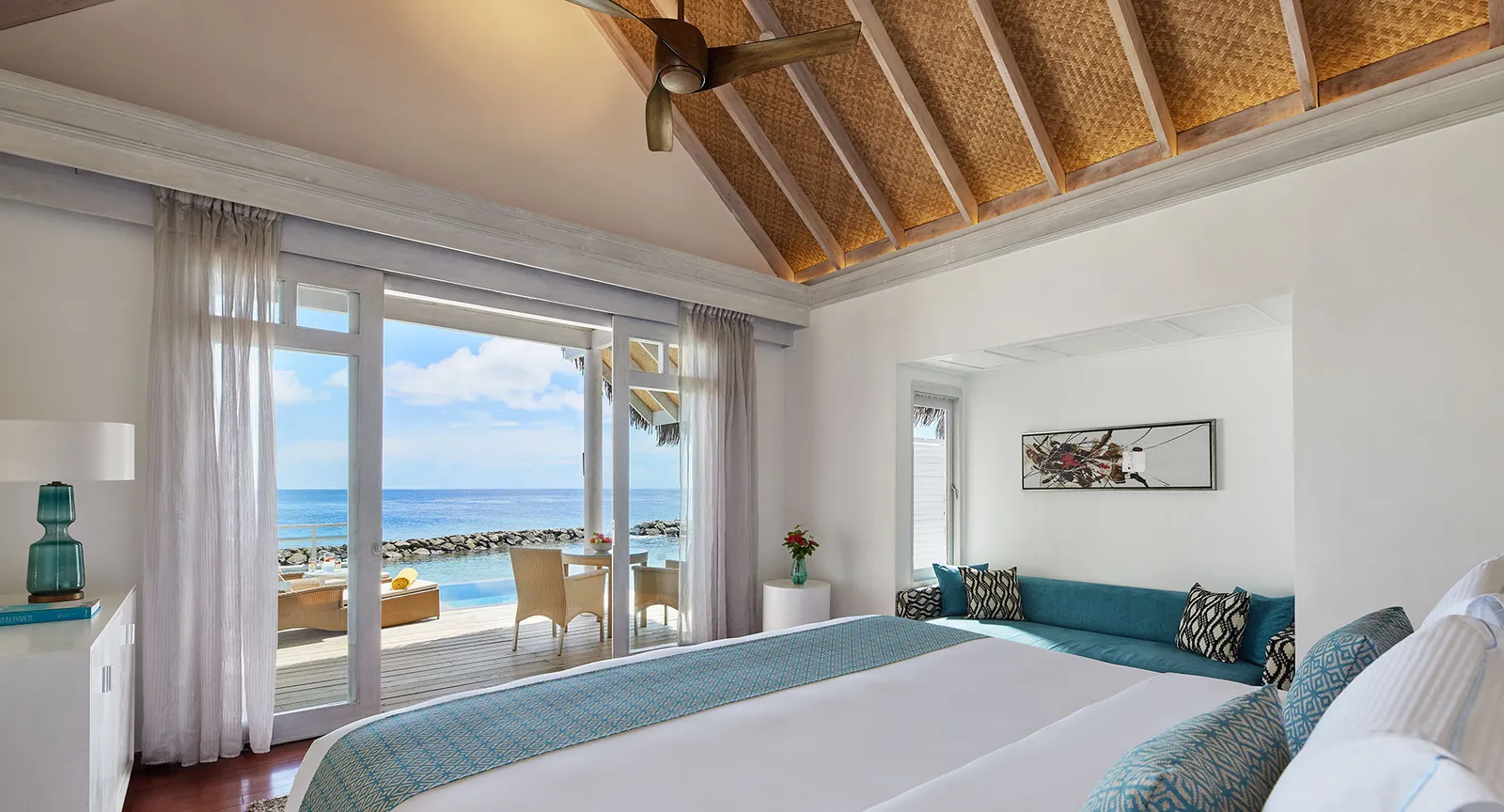 A water villa bedroom: the king bed, the timber ceiling and the glass doors open onto the deck and the sea.