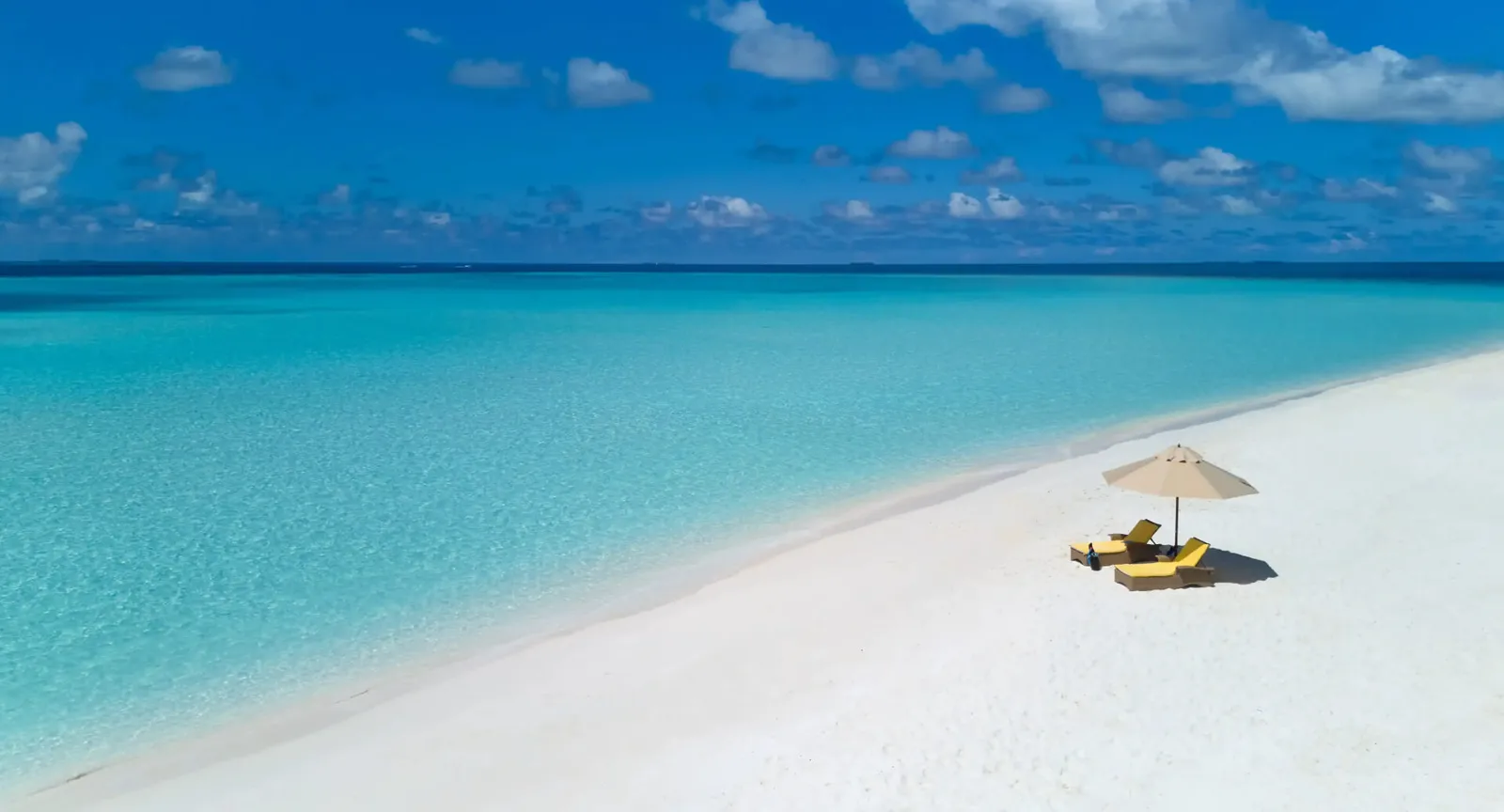 A parasol and 2 loungers on a white sandbank, between turquoise water and sky.