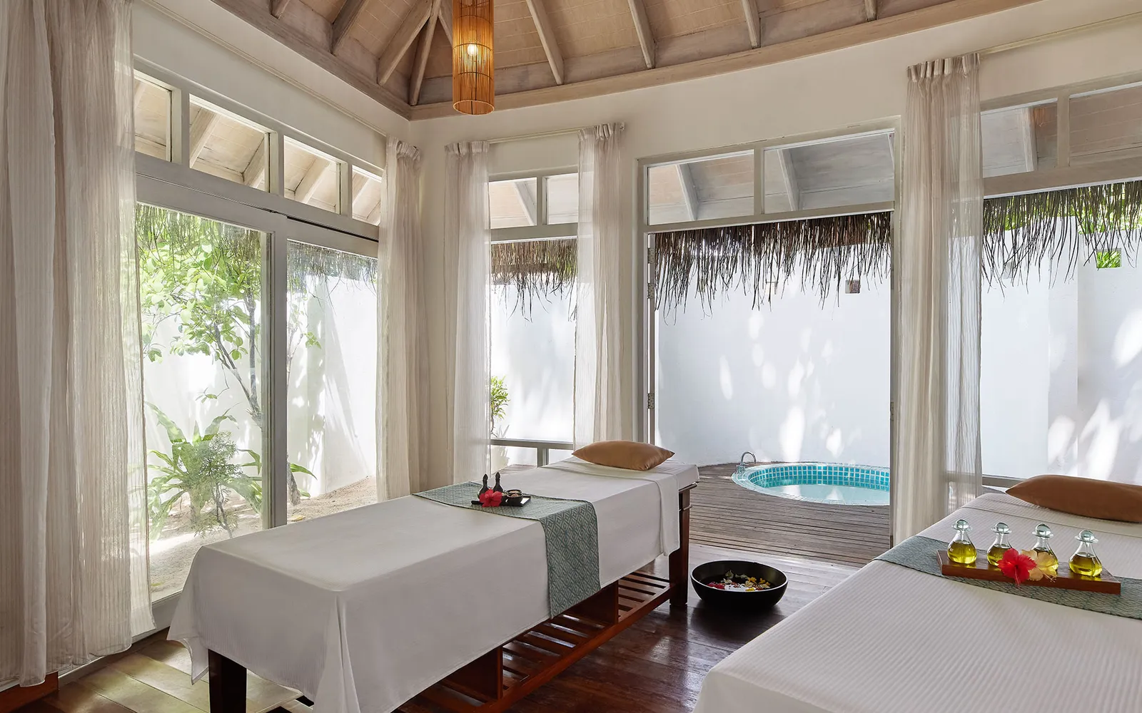 A spa room with 2 massage beds, white curtains and a whirlpool tub in the patio.