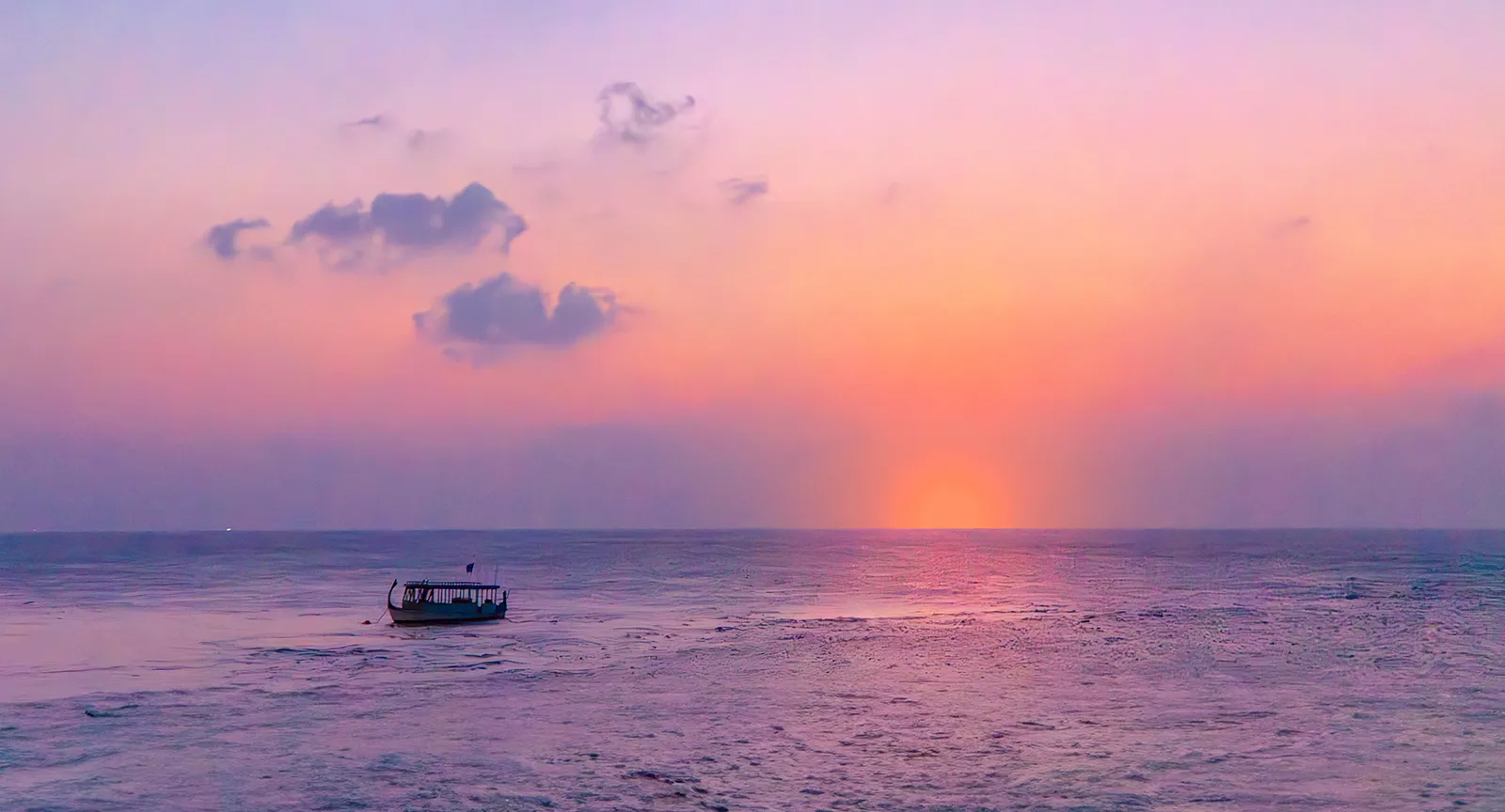 A Maldivian boat on the sea at sunset, under a pink and violet sky.