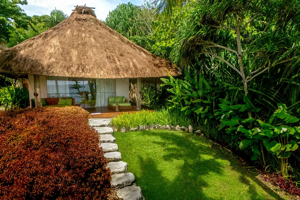 La Kasambi Suite: il bungalow dal tetto di paglia in fondo a un prato, con un sentiero di pietre fra le piante